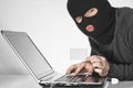 Hacker in balaclava holding a card in left hand and typing something with right hand on laptop keyboard Royalty Free Stock Photo