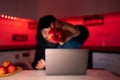 A hacker in a black hoodie with a hood on his head is sitting at a laptop holding a red apple in front of him Royalty Free Stock Photo