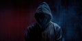 Hacker in black hoodie stands in front of dark blue background. Hood covers face, creating mysterious appearance
