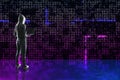 Hacker in hoodie holding laptop and standing on abstract glowing pixel wall background. Large led projection screens and data