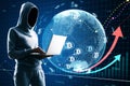 Hacker in hoodie using laptop with glowing globe hologram with financial arrows and money signs on blurry background. Digital Royalty Free Stock Photo