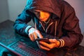 Hacker with phone is typing on a laptop keyboard in a dark room under a neon light. Cybercrime fraud and identity theft Royalty Free Stock Photo