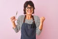 Hairdresser woman wearing apron and glasses holding scissors over isolated pink background screaming proud and celebrating victory Royalty Free Stock Photo