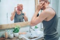 Hairless man touching his new style bald head haircut he made using an electric rechargeable Trimmer looking in bathroom mirror.