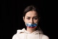 Half-legth portrait of young emotional upset girl with three colors duct tape over her mouth isolated on dark background