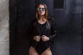 Half-length portrait of young woman wearing sunglasses, black swimsuit and leather jacket posing over dark