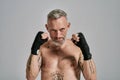 Half naked middle aged athletic man, kickboxer looking angry holding hands standing in studio over grey background. Muay
