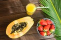 Half of Ripe Papaya Fresh Strawberries in Bowl Pineapple Citrus Juice in Tall Glass with Straw Palm Leaf on Plank Wood Table