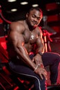 Halfnaked focused african american bodybuilder sitting in modern dark gym with red light.