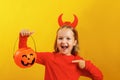 Halloween celebration. Little girl in a devil costume on a yellow background. The child holds a pumpkin jack lantern