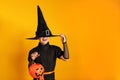 Halloween celebration. Little girl in a witch costume holds a pumpkin jack bucket on a yellow background. The child hid his face