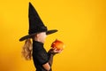 Halloween celebration. Side view of a little girl dressed in a witch costume holds a pumpkin on a yellow background Royalty Free Stock Photo