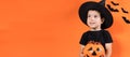 Halloween child. A preschool boy in a witch costume holds a pumpkin with candy on an orange background. Banner place for