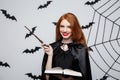 Halloween Concept - Beautiful Witch playing with magic stick and magic book on grey background. Royalty Free Stock Photo