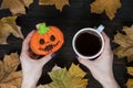 Halloween cookies jack o lantern and Cup of coffee in female hands. Background of autumn leaves Royalty Free Stock Photo