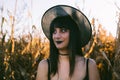 Halloween costume witch girl portrait in a cornfield at sunset. Beautiful serious young woman in witches hat with long black hair Royalty Free Stock Photo