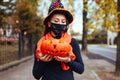 Halloween jack-o-lantern pumpkin. Girl holds carved pumpkin outdoors wearing mask against coronavirus.