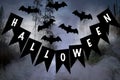 Halloween lettering on black flags. Nearby are figures of bats. Behind the forest and smoke.