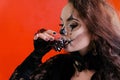 Halloween makeup. A brunette girl with long dark hair in a black dress drinks a bloody liquid from a glass in the form of a skull Royalty Free Stock Photo