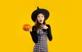 Halloween theme, young asian woman in black dress wearing witch hat holding orange pumpkin posing surprised and excited on yellow Royalty Free Stock Photo