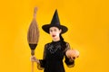 Halloween theme, young asian woman in black dress witch costume wearing black witch hat holding broom and pumpkin posing on yellow Royalty Free Stock Photo