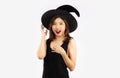 Halloween theme, young pretty asian woman in black dress wearing witch hat posing surprised and wow on white background Royalty Free Stock Photo