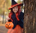 Halloween trick or treat.Happy girl wearing a witch`s hat, with a pumpkin handbag, peeps out from behind a tree.. Funny kid in Royalty Free Stock Photo