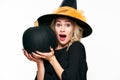 Halloween Witch with shocked expression holding large black pumpkin. Beautiful young woman in witch hat holding pumpkin. Royalty Free Stock Photo