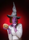 Halloween. A young woman with purple skin in a black witch hat is holding a pumpkin with a zombie face. Dark red background. Royalty Free Stock Photo