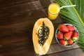 Halved Ripe Papaya Fresh Strawberries in Bowl Pineapple Juice in Tall Glass with Straw Palm Leaf on Plank Wood Table in Cafe