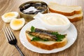 Halves of eggs, opened jar with sprats, slices of bread, fork, sandwich with sprats, bowl with mayonnaise in plate on table