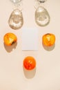 Halves of persimmon fruit with white paper card note and two glasse with water on beige pastel background. Summer refreshment