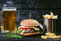 Hamburger with french fries, beer on a burnt, black wooden table. Fast food meal. Homemade hamburger consist of beef meat, lettuce