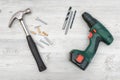 A hammer, a cordless drill with a set of bits, several bolts, dowels and plugs on white wooden background.