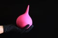 A hand in a black glove holds a pink enema on a dark background