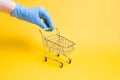 a hand in a blue disposable rubber glove holds a miniature shopping trolley by the handle, shopping during a pandemic