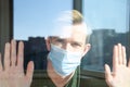 Hand of caucasian man in medical mask on glass of window on self-isolation during an epidemic. Blurry, defocus, light reflection.