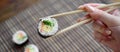 A hand with chopsticks holds a sushi roll on a bamboo straw serwing mat background. Traditional Asian food
