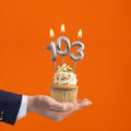 The hand that delivers cupcake with the number 103 candle - Birthday on orange background