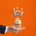 The hand that delivers cupcake with the number 107 candle - Birthday on orange background