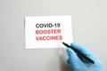 The hand of the doctor with the blue glove writes on white paper the text `Covid-19 Booster Vaccines`. Concept of Combating the