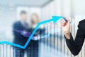 A hand is drawing a growing arrow on the glass scree, Blue dark background with financial graphs.