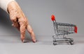 Hand of elderly woman approaches an empty small shopping cart. Gray background. The concept of internet shopping