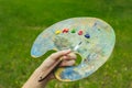 The hand of a European artist holds a mastekhin smeared in oil paint and a palette against the background of a green lawn. Blurred Royalty Free Stock Photo