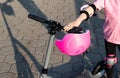 The hand of a girl in protective gear is holding the steering wheel of a scooter on which a protective pink helmet hangs