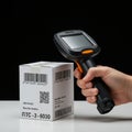 A hand-held barcode scanner is positioned near a white box featuring multiple barcodes and a QR code.