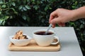 Hand hold spoon stirring coffee on wood tray and there is cookie at side. Table is set in garden with green leaves background. Royalty Free Stock Photo