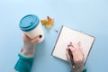 Hand holding bamboo reusable takeaway cup with lid on and writing on blank pages of notebook, on blue background