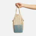 A hand is holding a beige, netted reusable shopping bag against a white background.