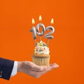 Hand holding birthday cupcake with number 102 candle - background orange